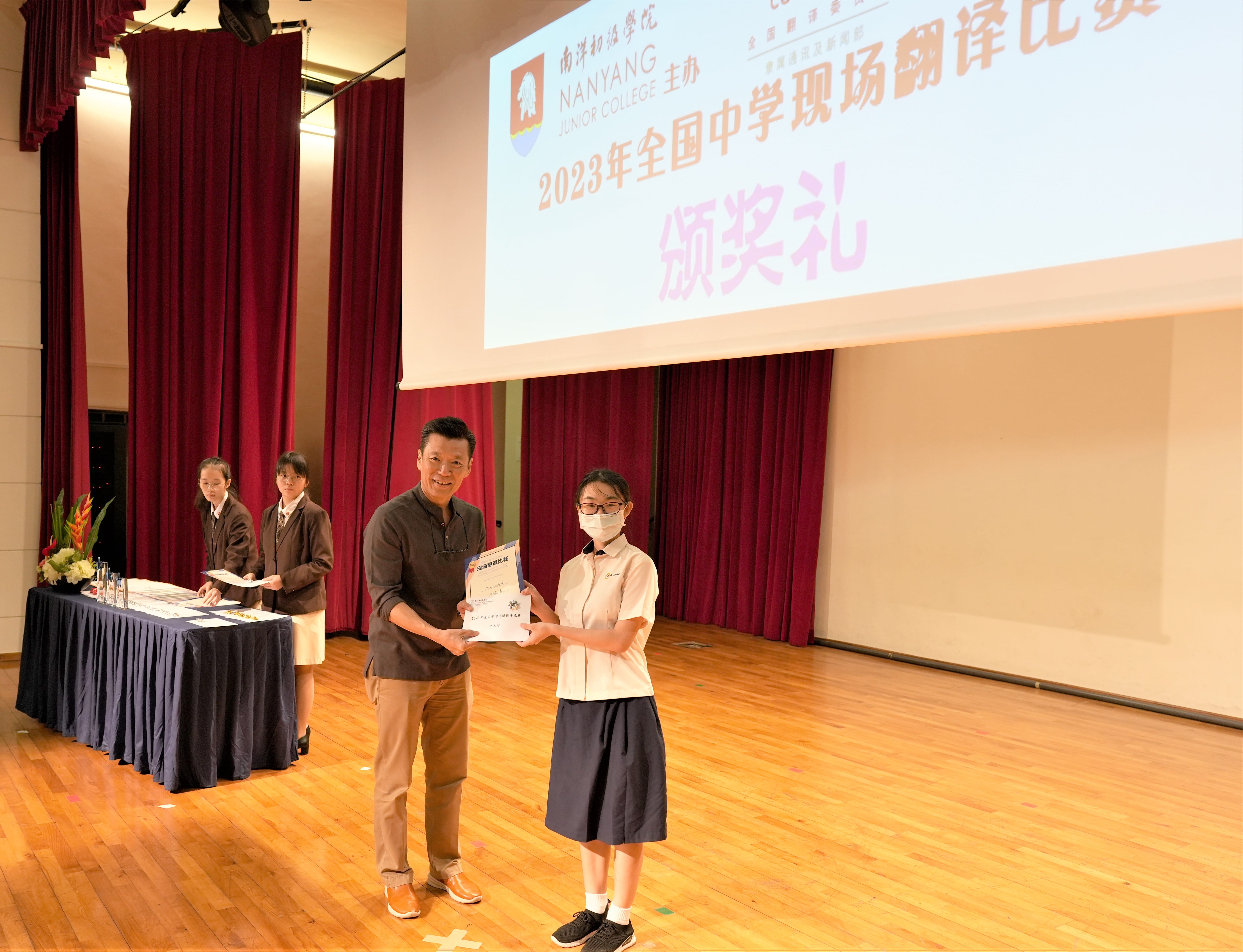 A man presents a certificate to a masked student on stage at a 2023 Nanyang Junior College award ceremony.